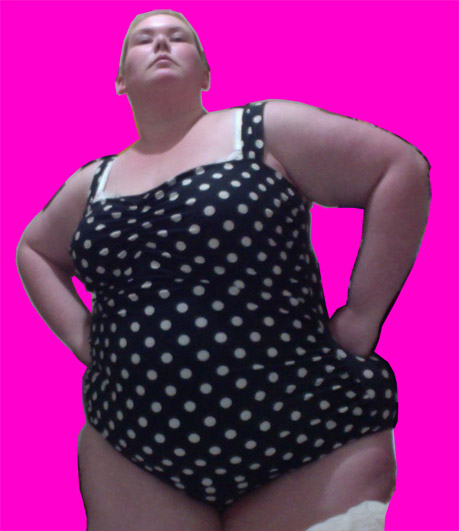 A photo of me, a fat white woman in a black and white polkadot one piece bathing suit, posing with hands on hips in front of a hot pink background.